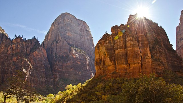 Sunshine peaking out behind a red sandstone cliff. A tall mountain is in the background.