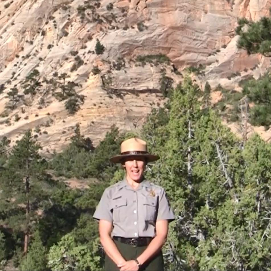 A ranger wearing a green and grey uniform and a flat hat stands before a forested sandstone cliff.