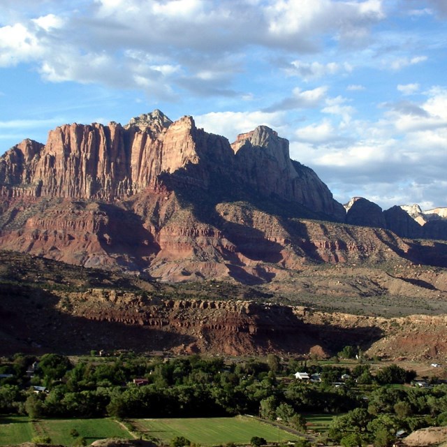 Red, orange, and beige sandstone cliffs toweing over green trees and the town of springdale