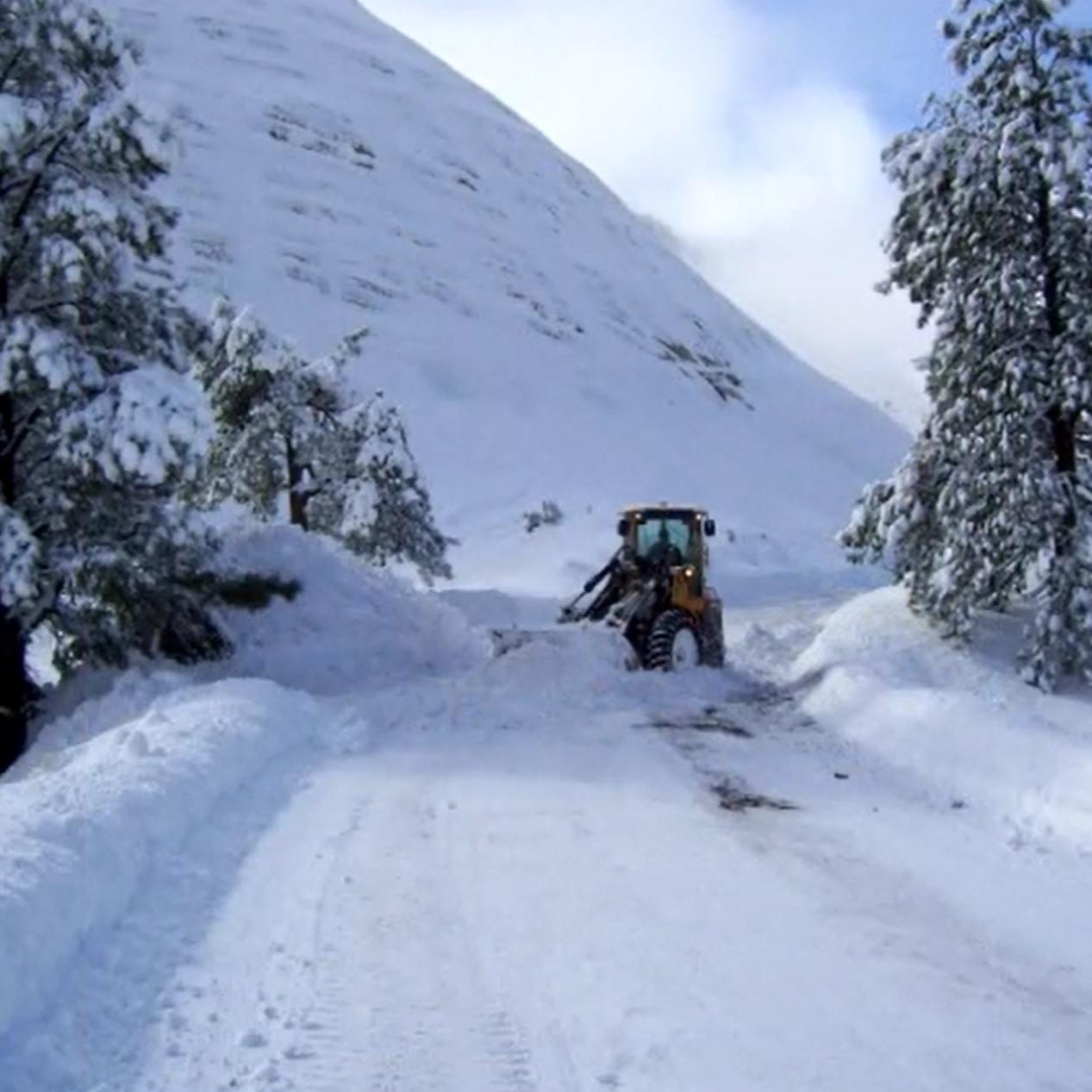 A snow plow clears over a foot of snow off of an impassable road. Snow-covered trees line the road.