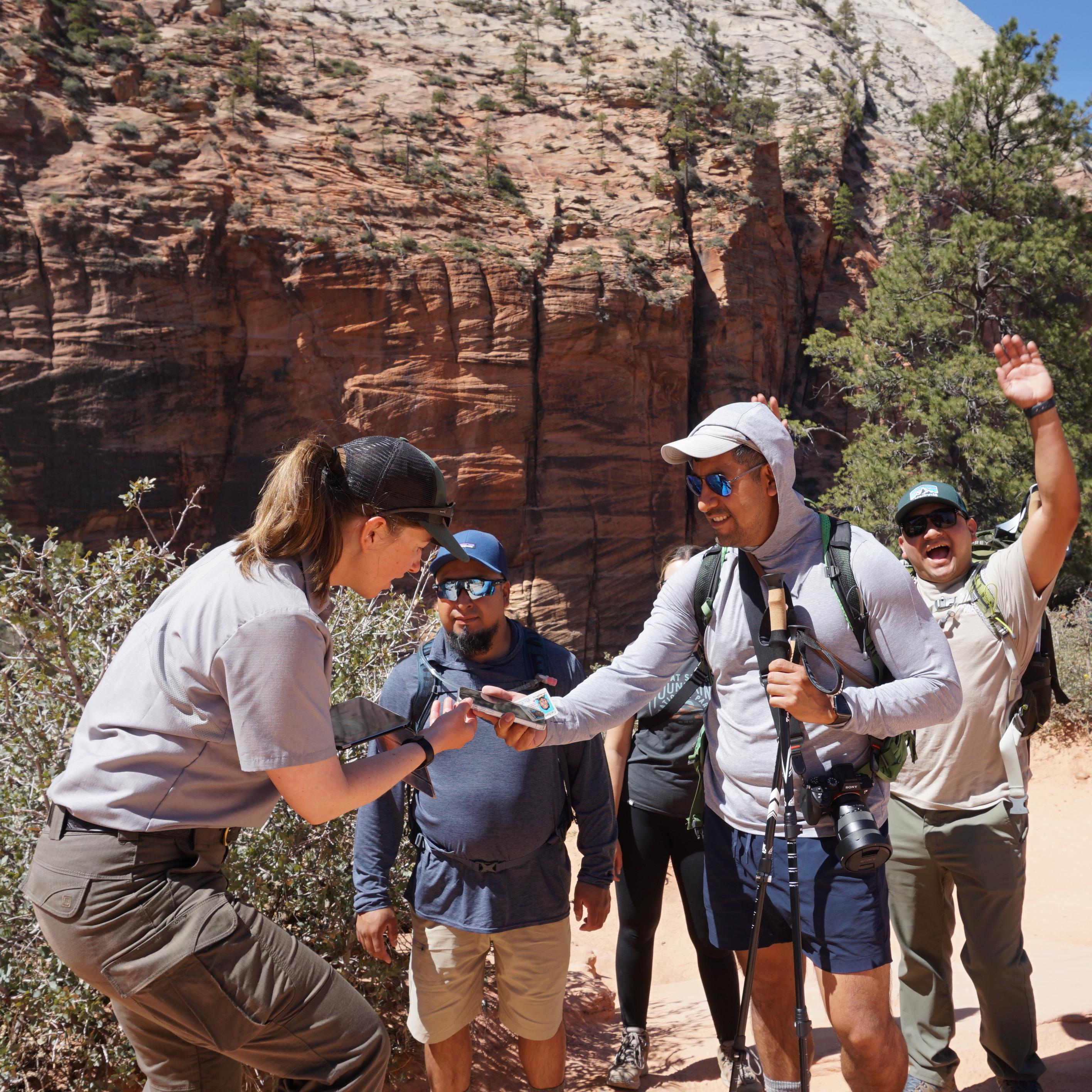 A ranger looks at a phone of a visitor next to a group with orange sandstone cliffs and a canyon