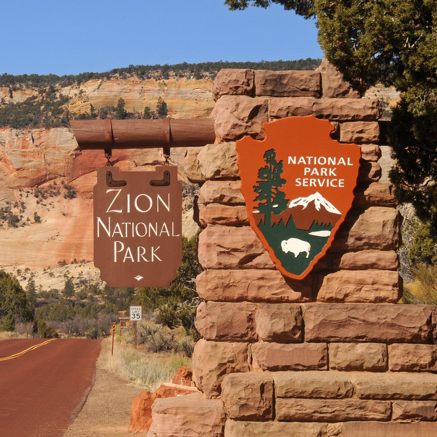 The Zion entrance sign, with a red road and red sandstone cliffs in the background. 