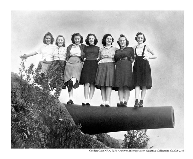 A black and white photo shows seven teenage girls playfully posing for a photo while standing on top
