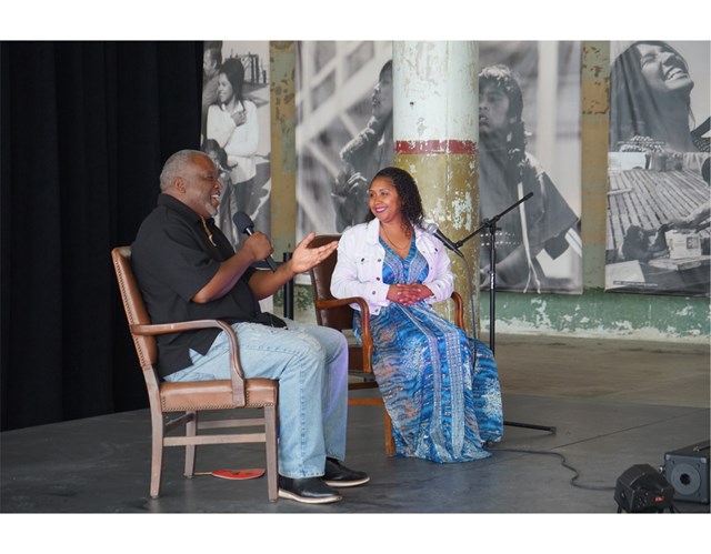 A woman and a man sit with microphones onstage, smiling, during a live event.