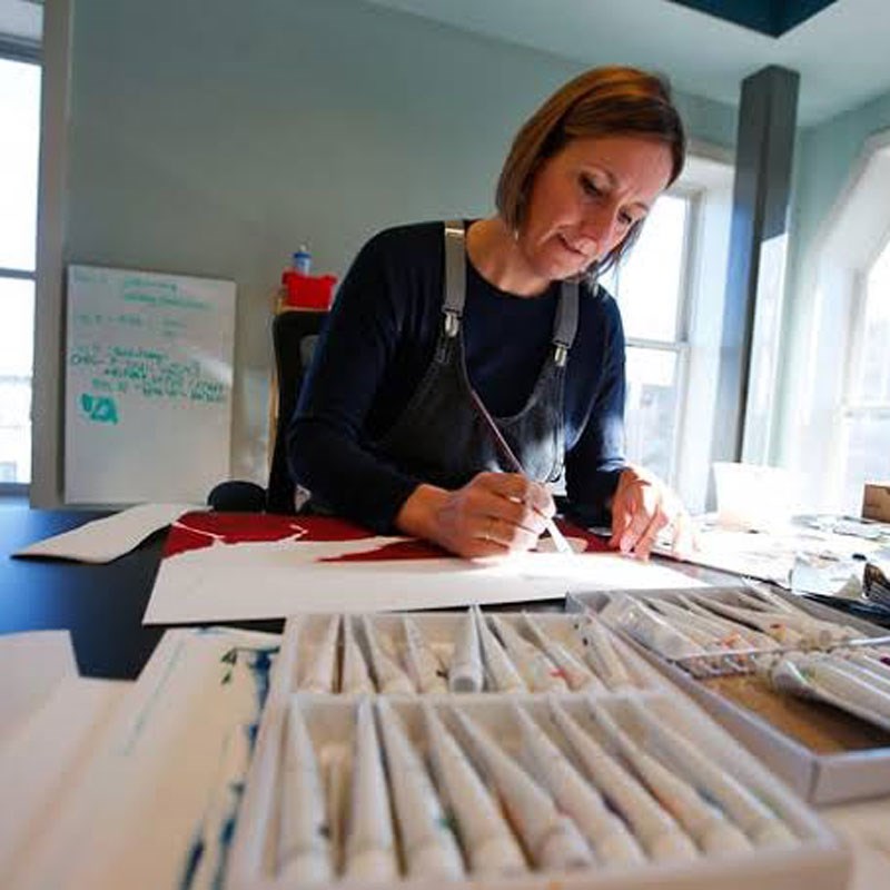 Margo sits at a table painting with watercolors, surrounded by natural light and supplies.