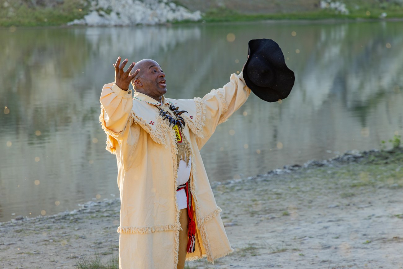 A man stands in front of a river, portraying York with arms outstretched, holding a black hat