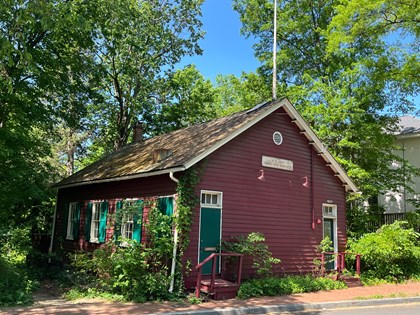 A dark red clapboard-sided building with two doors on the front. Each door has a couple of steps leading up to it. The doors are green, each with a three-pane transom window above it. A sign with the name of the building is high on the exterior wall, above and about halfway between the doors. Along the side of the building are three large windows with green shutters. Vines are growing up along the side of the building in some places.