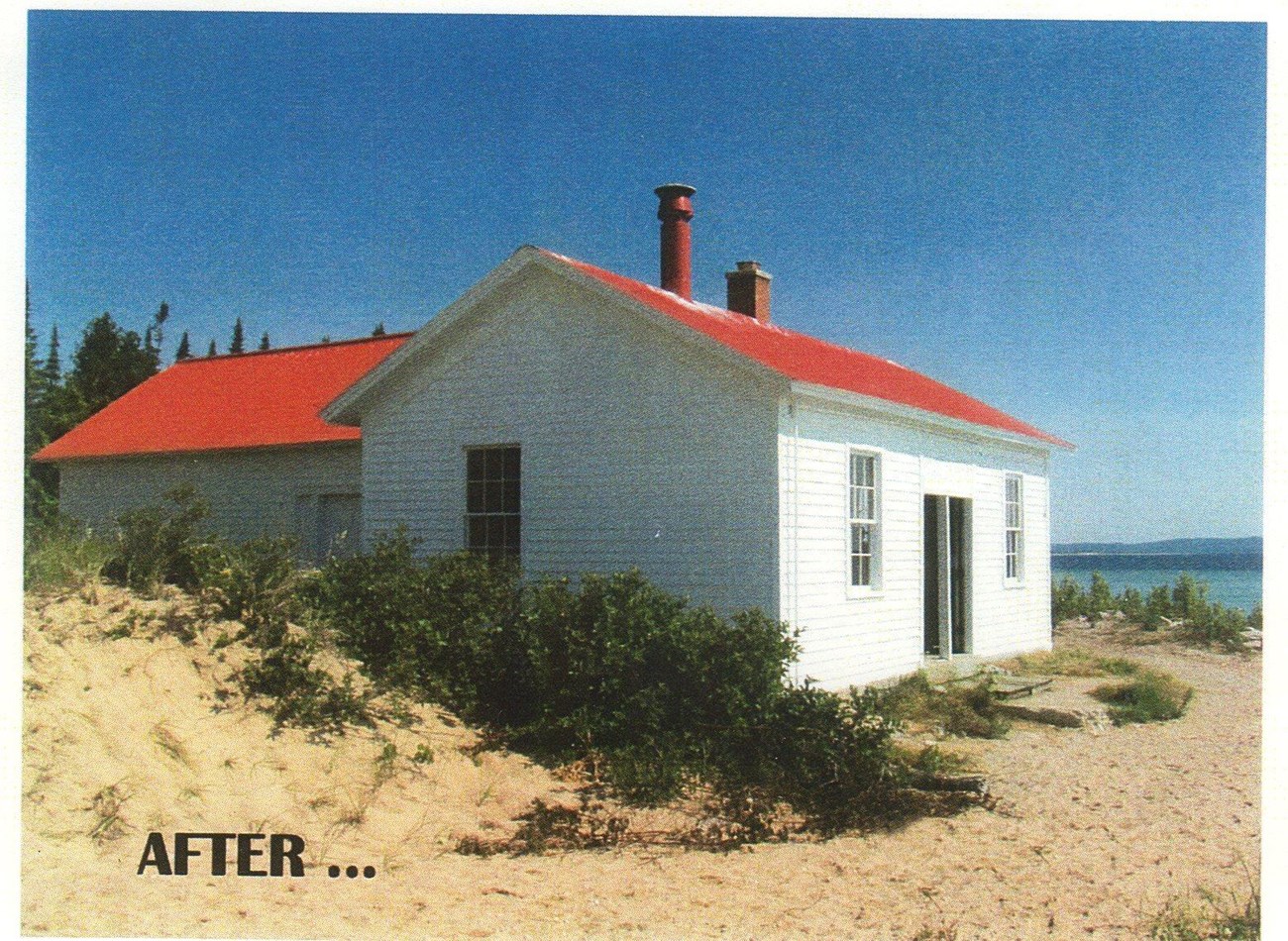 A T shaped white building with a bright red roof surrounded by a sandy beach.