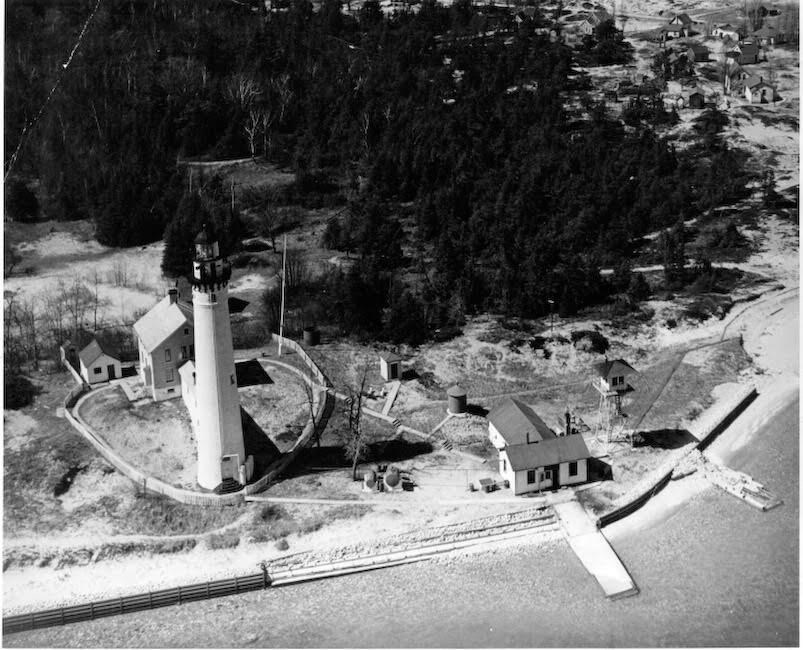 A historic black and white aerial photograph of several buildings and a lighthouse along a sandy lakefront beach.