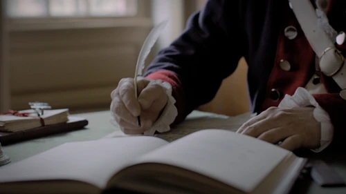 a light skinned man writes with a quill pen in an open book