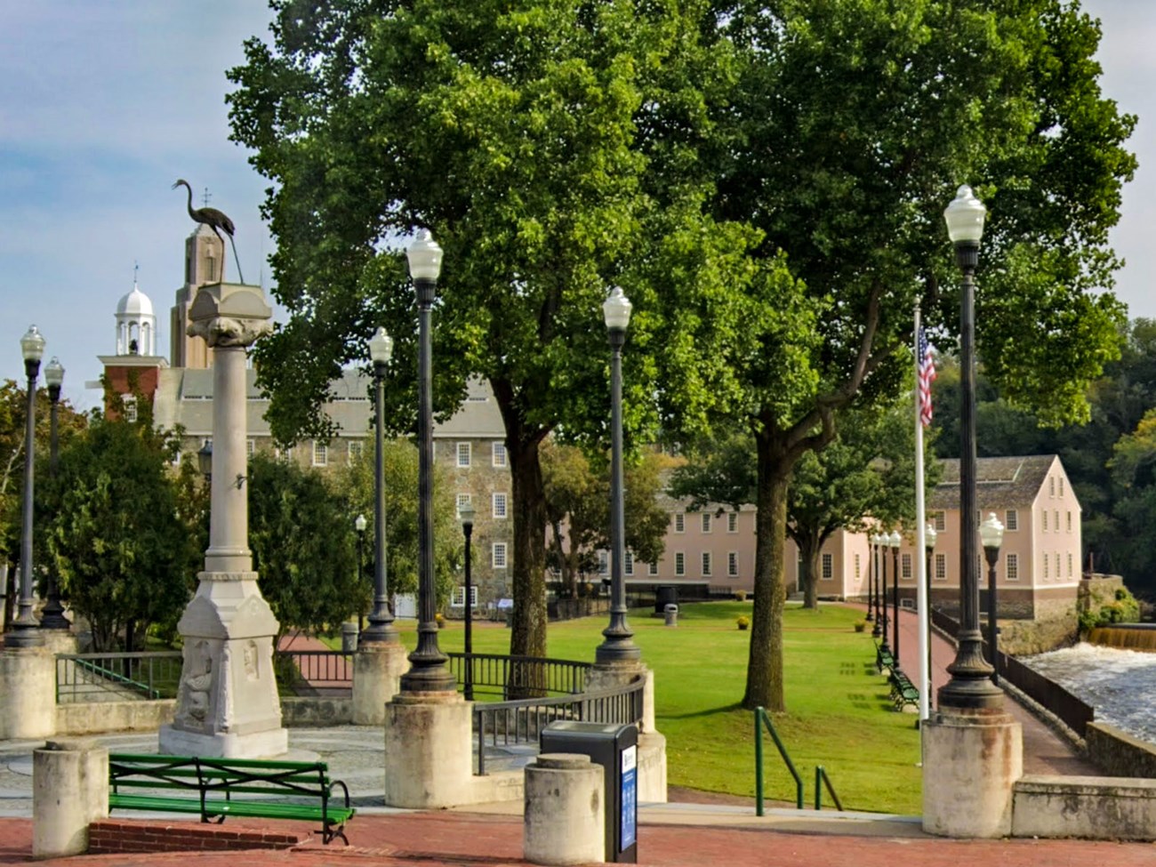 A riverside park with a monument, lampposts, and historic mill buildings in the background.
