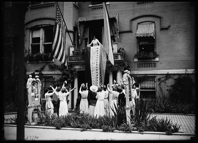 A group of woman cheering below a balcony where a woman drapes down a flag toward them.