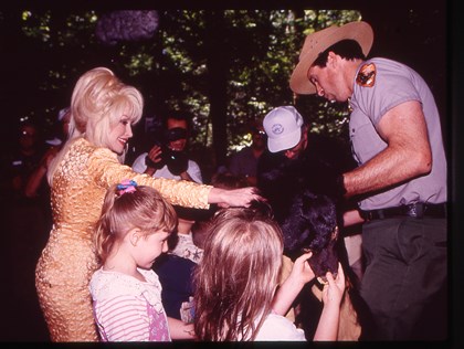 Dolly Parton in a yellow sparkly dress petting a bear pelt amongst many children and a ranger
