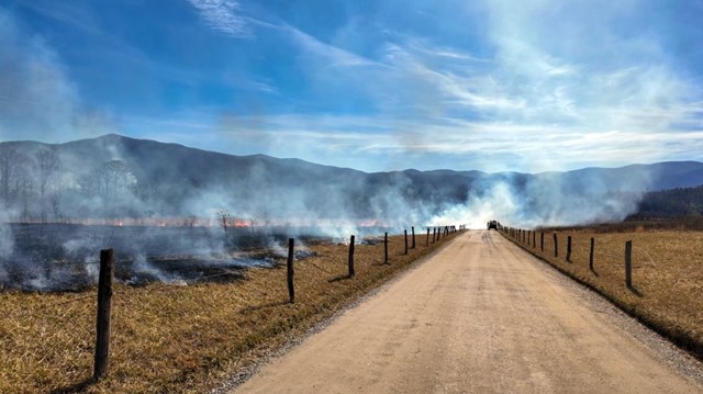 Smoke lingering over a dirt road with fields on either side and an ATV in the distance