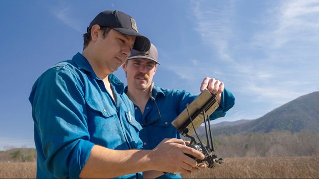 Two people looking at a tablet that is controlling a drone with mountains and a blue sky behind them