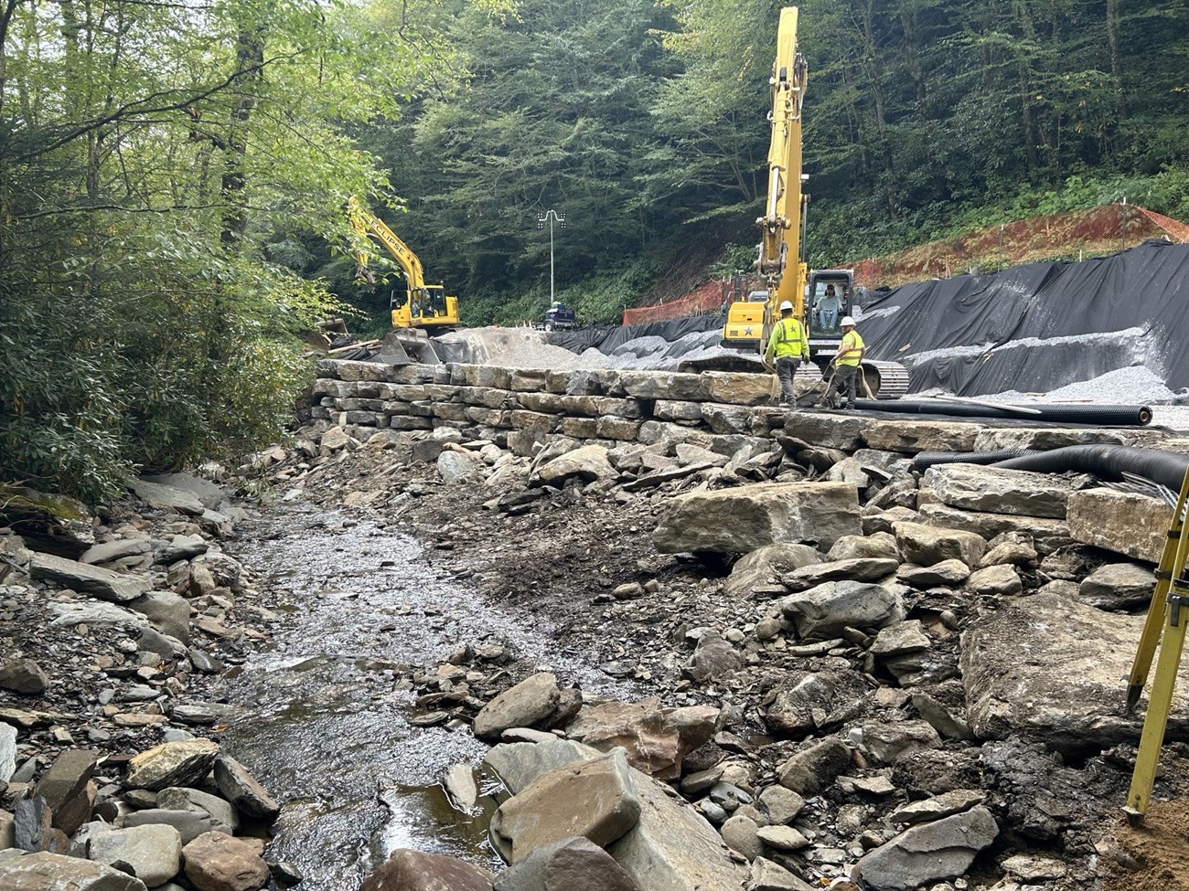 Crew building a stone retaining wall along a roadway and stream.