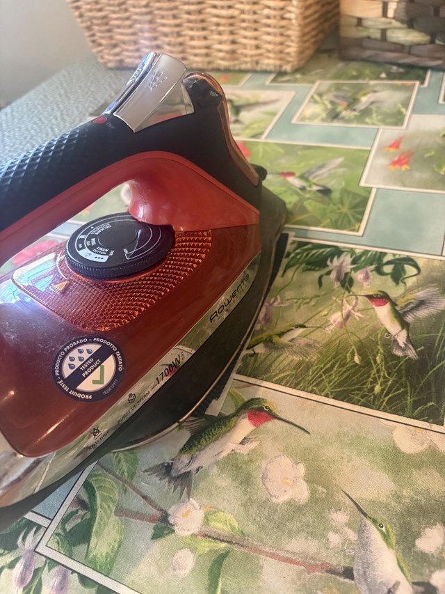 A red iron on an ironing board with a green hummingbird cover.