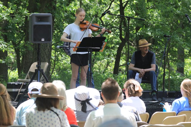 A young woman stands on an outdoor stage playing a fiddle. A man sits on stage behind her and the audience watches attentively.