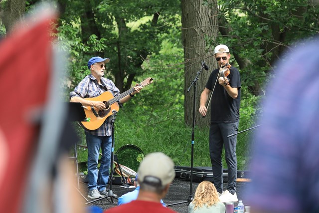 Two men stand on an outdoor stage playing stringed instruments. The one on the left plays the guitar, and the one on the right plays the fiddle. Audience members are seen watching in the foreground.