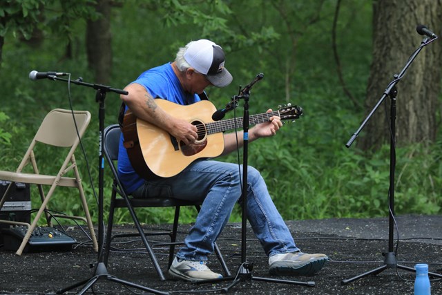 White haired man dressed in blue shirt and jeans and wearing a ball cap plays guitar on an outdoor stage.