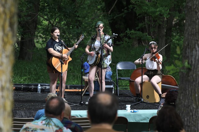 Three young women play stringed instruments on an outdoor stage. Two of the women appear to also be singing.