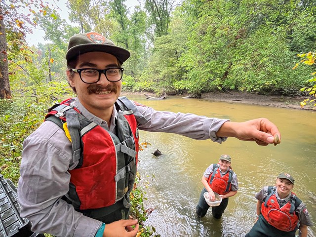 Person in outdoor gear holds a small mussel by a river while two others stand in the water below.