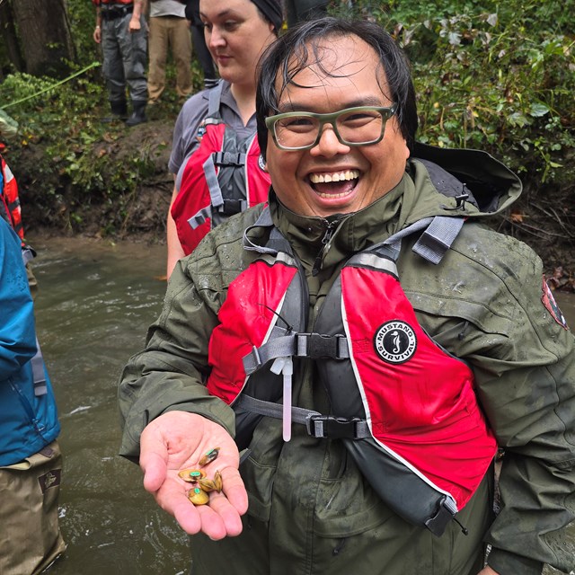 Person in outdoor gear holds small mussels by a creek while standing with a group.