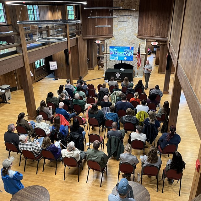 Audience seated in a wood‑paneled hall listens to a presenter beside a screen and podium.