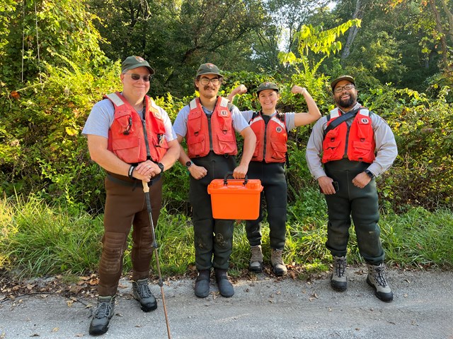 Group wearing outdoor gear and red vests stands by greenery with one person holding an orange case.