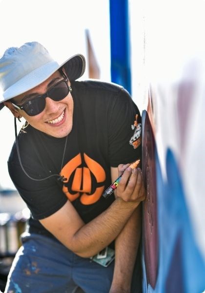 A person leaning against a wall, smiling at the camera, with a paint marker in hand