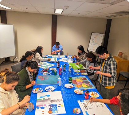 A group of people sitting at a table, individually painting canvases