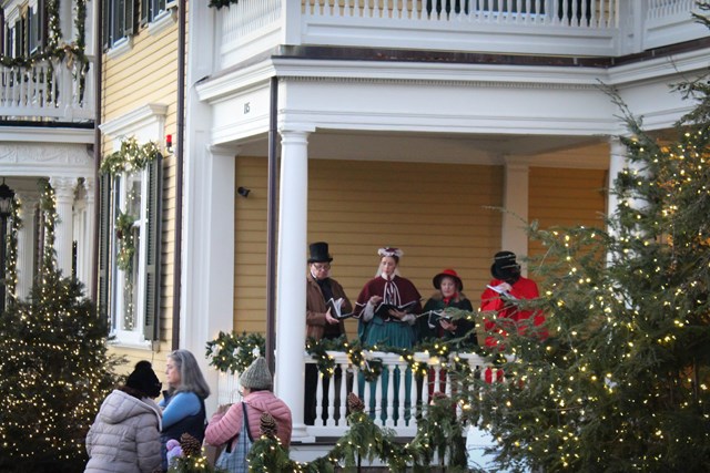 Carolers in Victorian attire sing on the porch of a historic house