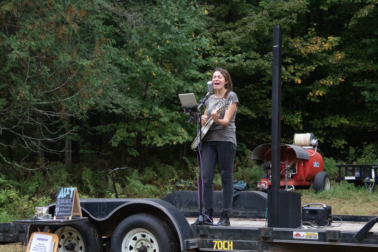 Woman sings into microphone on stand holding guitar while standing on a makeshift stage made from a trailer