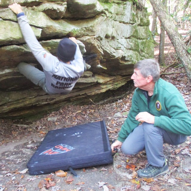 A man holding a drop pad under a rock climber bouldering.