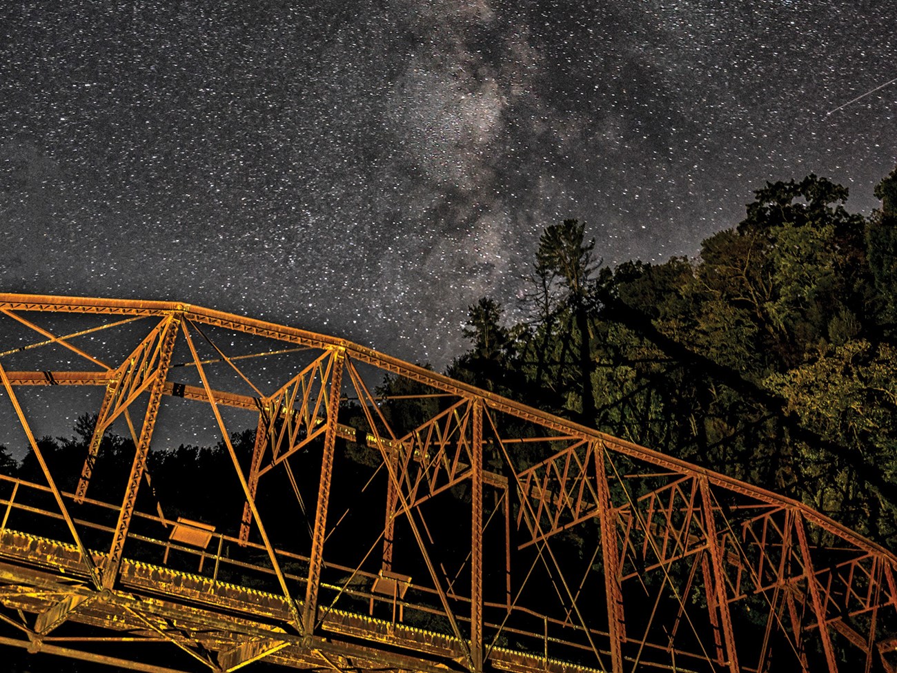 A metal bridge situated in front of a backdrop of trees, along with numerous stars and the Milky Way