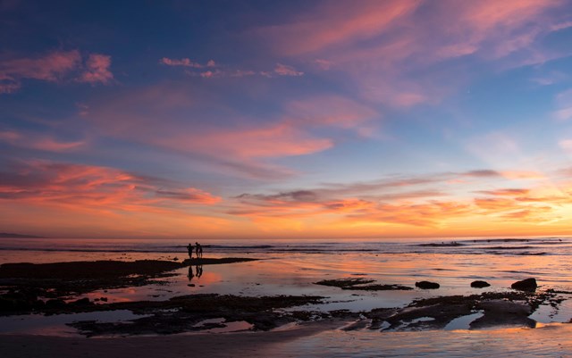 Sky and clouds in brilliant shades of yellow, orange, pink, and blue, reflected in calm tidepools. Two people are sillouhetted on some rocks towards the center left of the frame.
