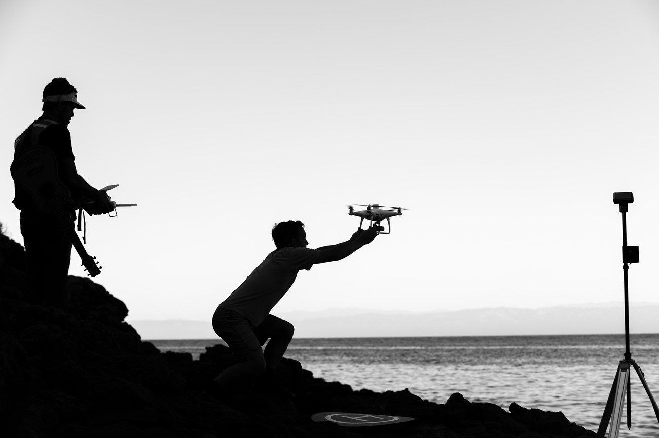 Sillouhettes of two people on a rocky bit of coast on a bright yet overcast day. One is operating a small drone controller with both hands as the other reaches out from a kneeling position, grabbing the drone from the air.