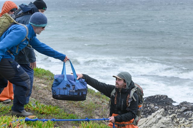 Researchers lean forward, handing an insulated bag to a colleague down a steep hill from them. The receiving researcher takes the bag with one hand and holds onto a knotted blue rope for stability with the other. A rocky shore and the sea are behind them.