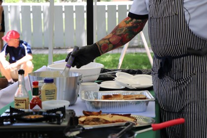 Chef with glove and a colorful tattoo sleeve of fruits and vegetables stirring a pot of cheese sauce