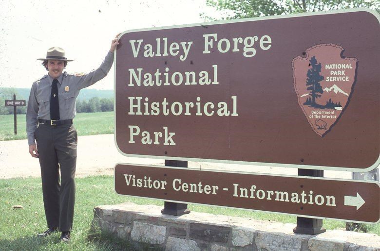 a park ranger stands in front of a large brown sign reading 