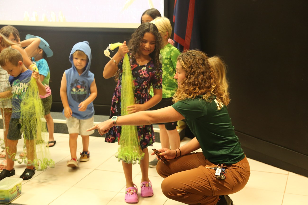 A curly-haired white woman crouches down to show a little girl how to use a fishing net.