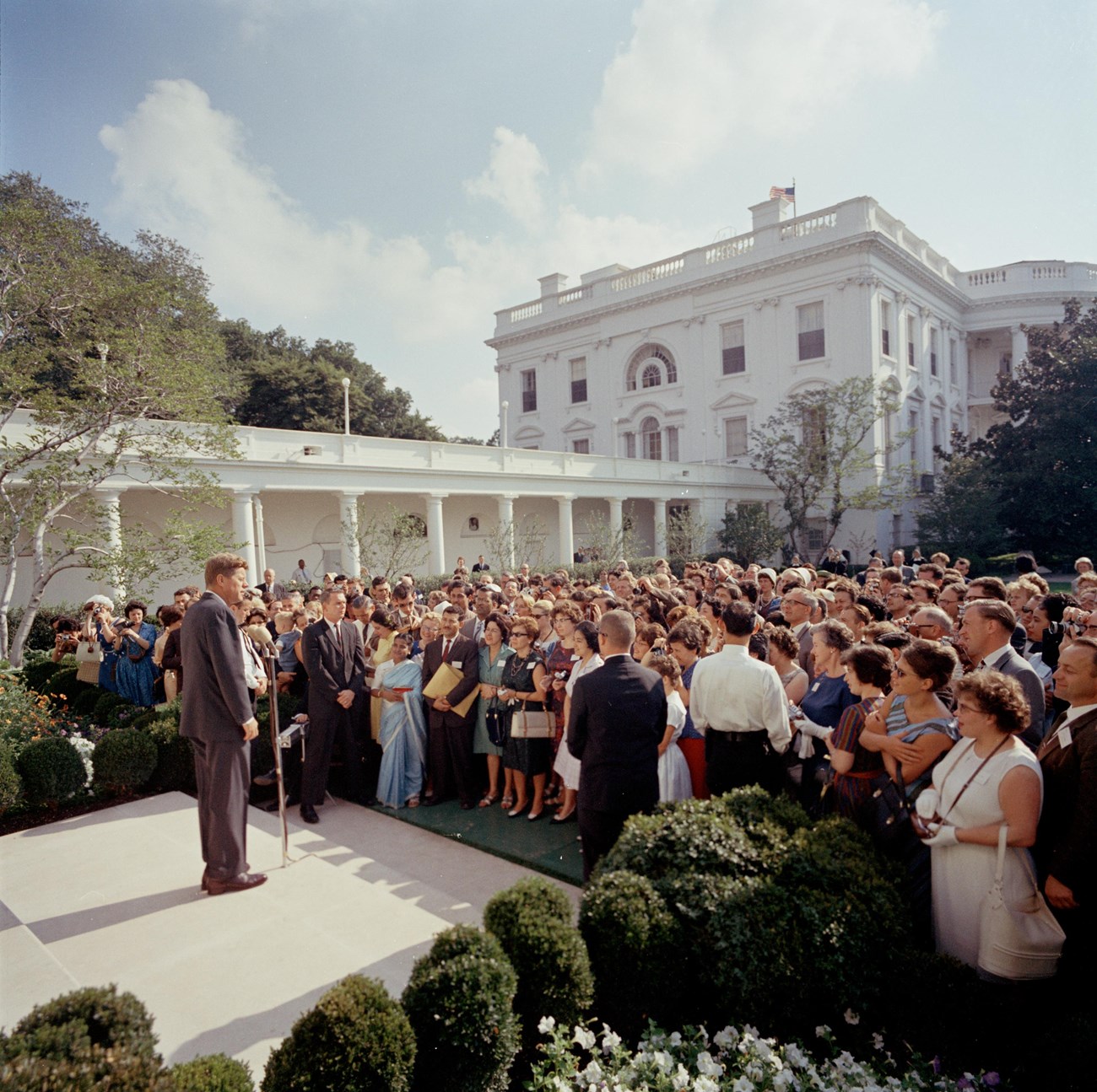 John F. Kennedy speaks before a crowd of people in the Rose Garden on a sunny day.