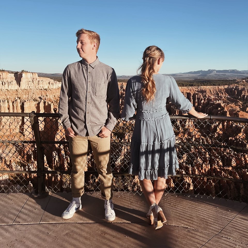 A couple in formal 1950s clothing stand at a railing overlooking a vast landscape of red rock spires and cliffs