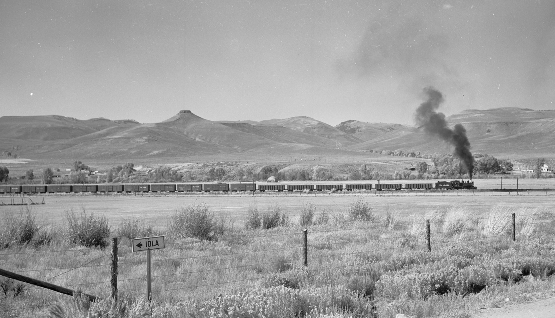 Historic black and white image of a train passing through a valley with mesas in the background