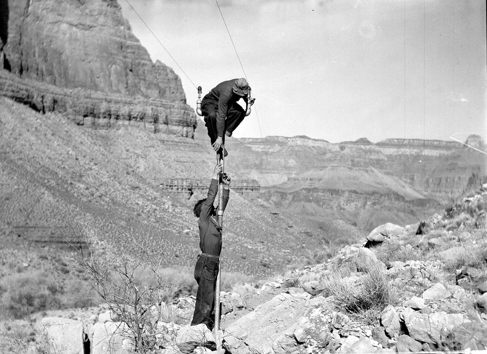 Two men, one atop a pole, the other holding it steady. Cliffs in background