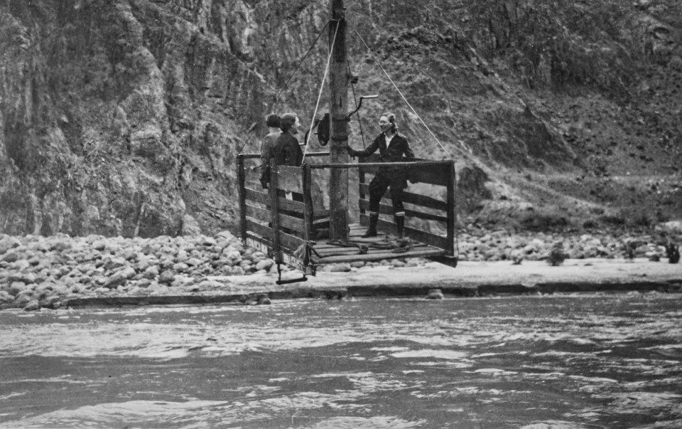 Women in a large wooden box above a river with cliffs in background