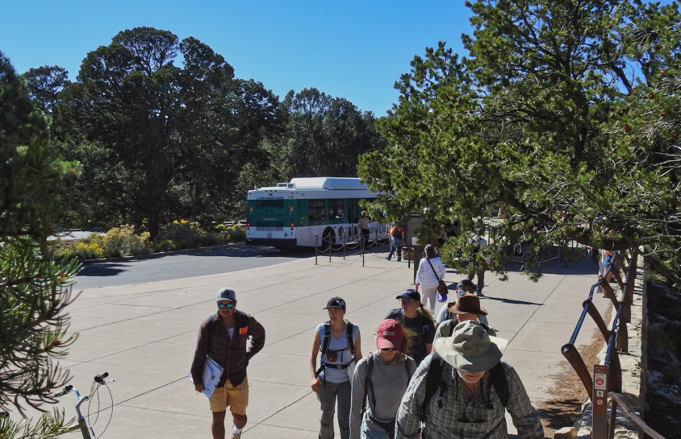 Multiple parked cars, people, a fence, road and trees