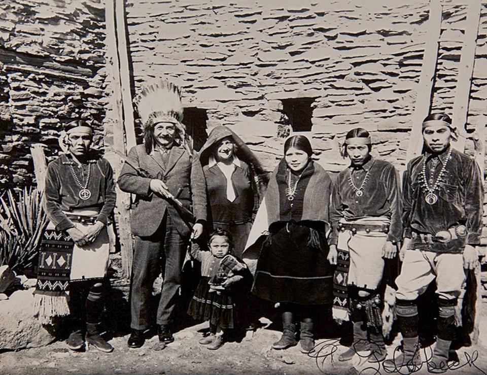6 people stand in front of stone building, most dressed in tribal clothing