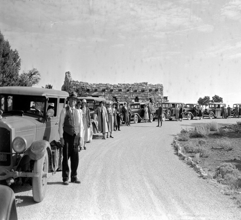 People and a line of old cars on road with stone building in background