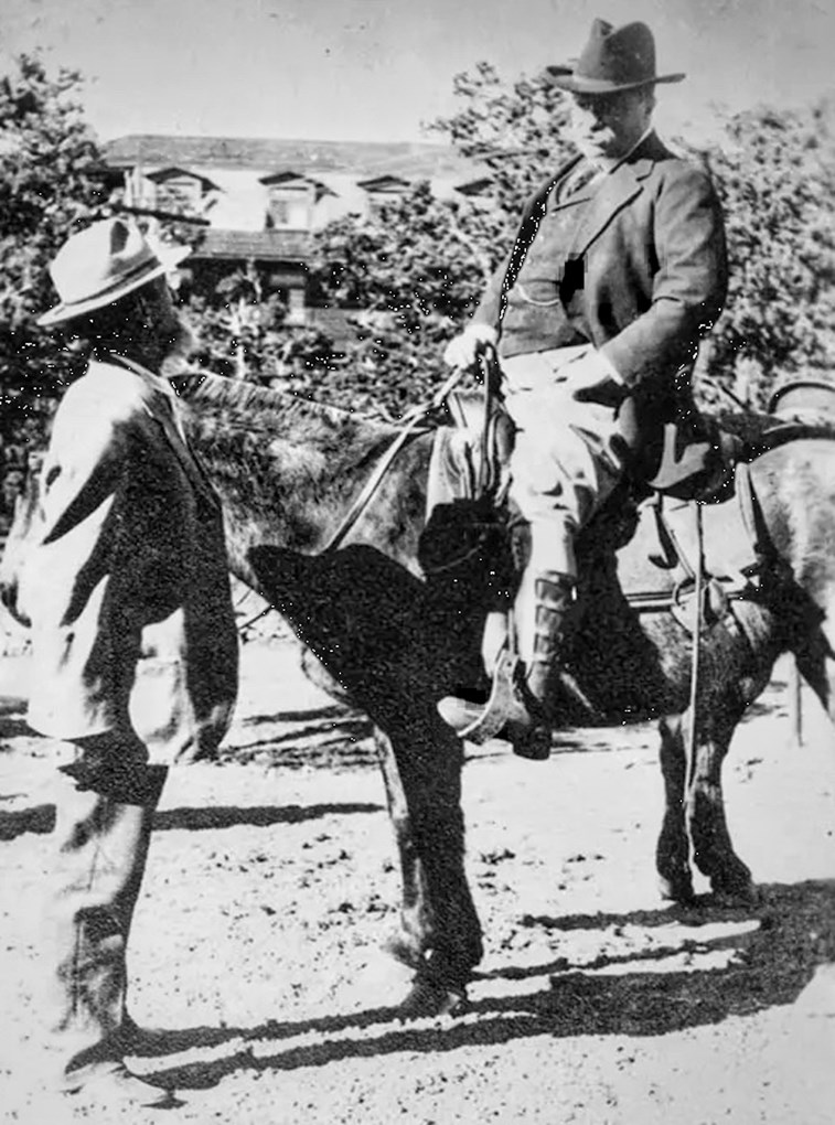 Man on horse with another man standing in front with buuilding and trees in background